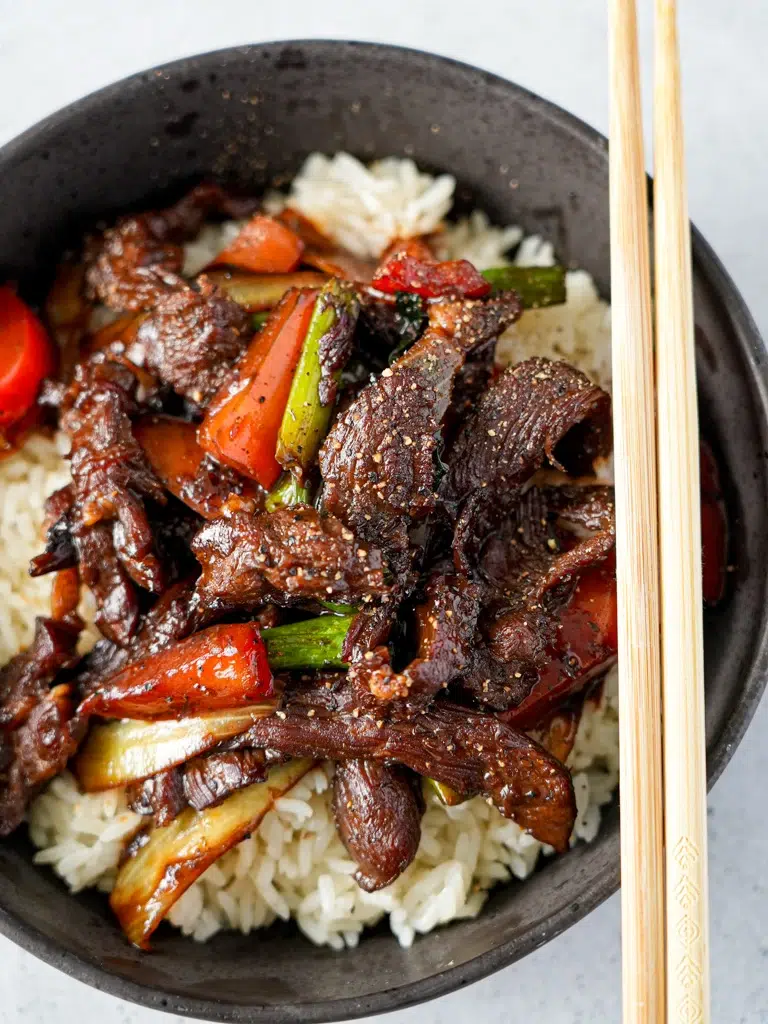 A bowl of black pepper beef stir-fry featuring tender beef slices, colorful bell peppers, and green onions, served in a rich black pepper sauce and chopsticks on side